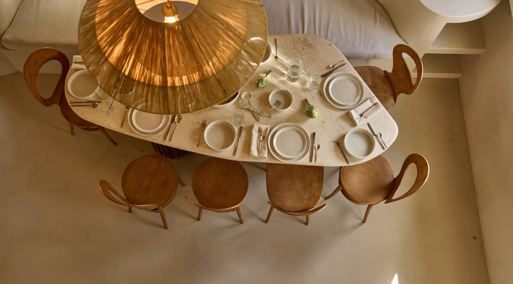 Ein unregelmäßig geformter Steintisch von oben. Darauf sind weiße Teller gedeckt. Den Tisch umrunden Holzstühle. Ein Teil der Deckenlampe ist zu erkennen.