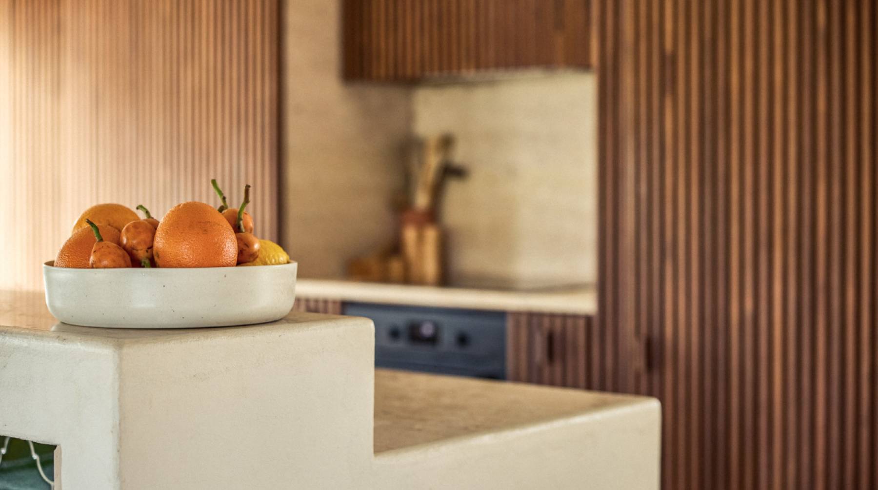 Hellbeige, ovale Keramikschale voller Orangen und kleiner Mandarinen steht auf einer cremefarbenen Trespenstufe; im unscharfen Hintergrund eine moderne Küche mit Holzlamellen-Schrankfronten und ein dunkler Ofen.
