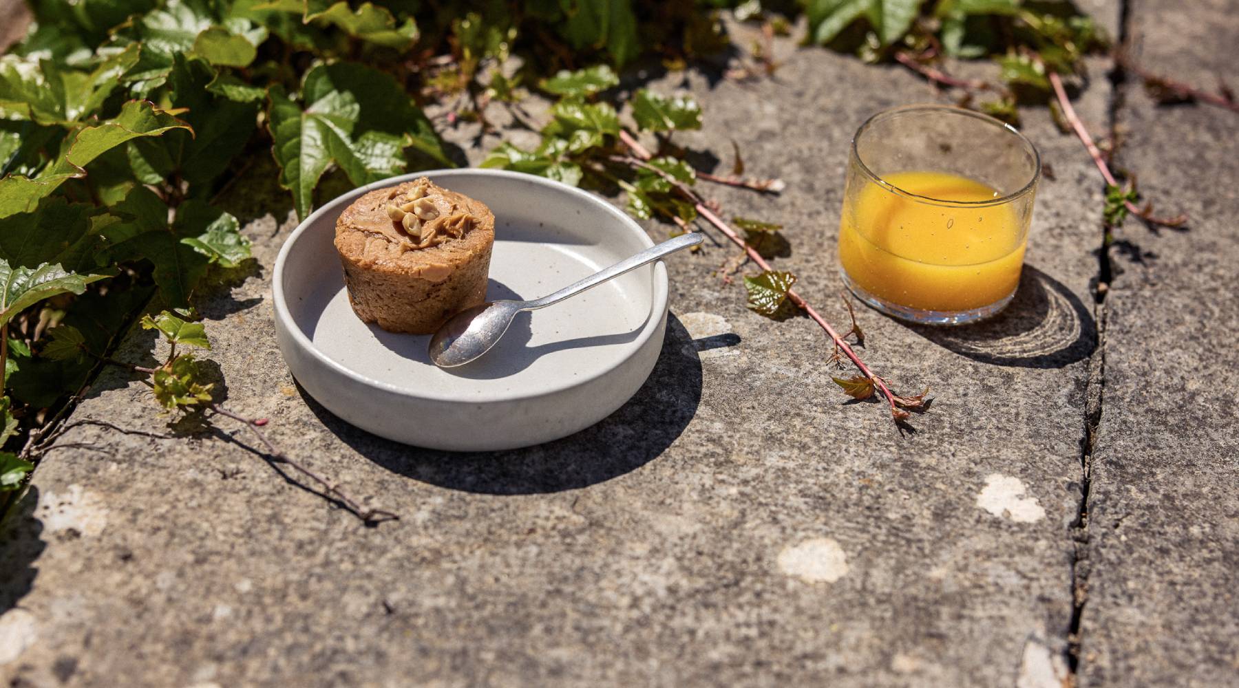 Auf einem Steinboden im Freien Steht ein kleiner Teller mit hohem Rand in Weiß. Darin ein kleiner Kuchen mit einem Löffel. Daneben steht ein Glas gefüllt mit Orangensaft.