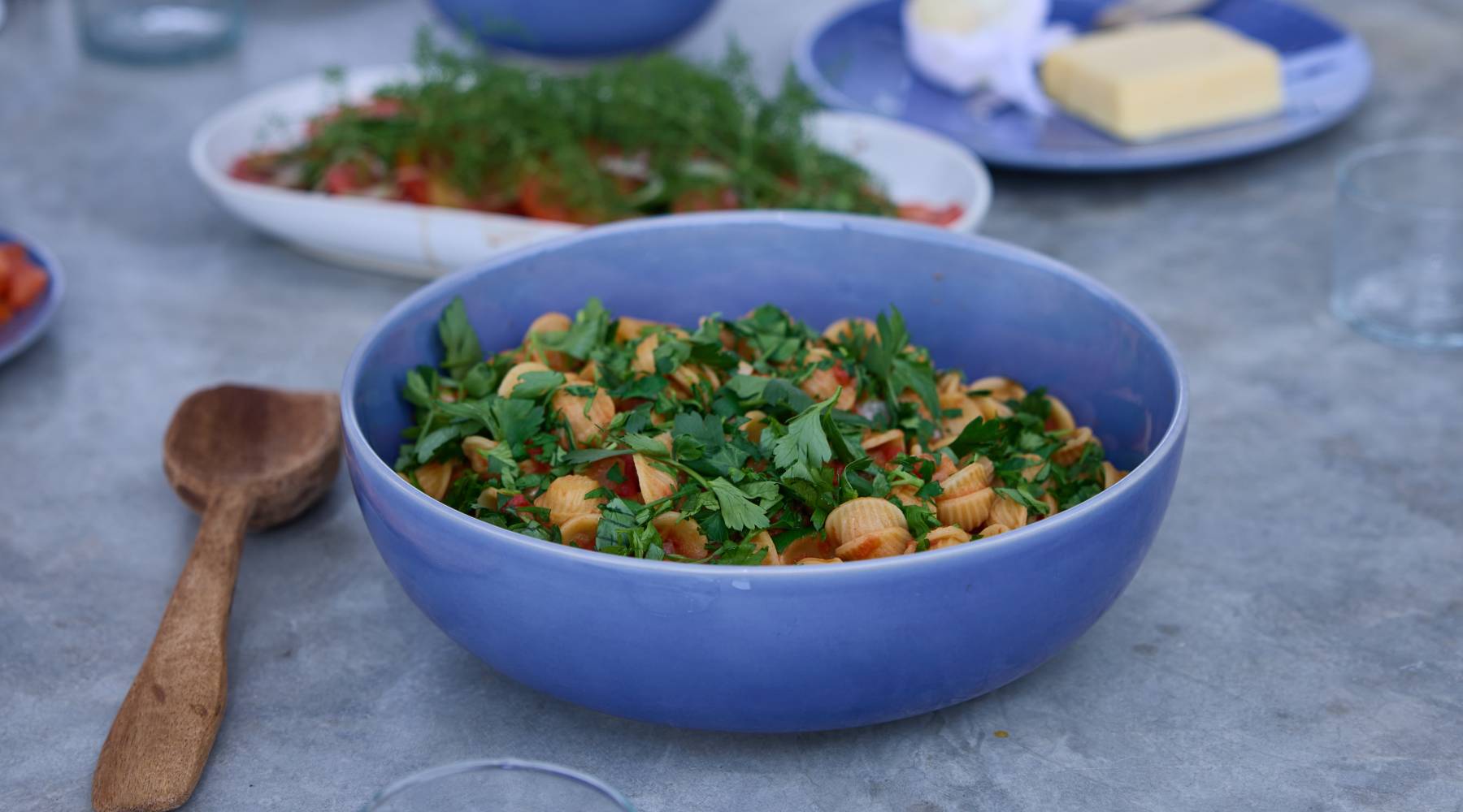 Große, matte blaue Keramikschüssel voller Muschelnudeln in Tomatensauce, großzügig mit gehackter Petersilie bestreut, steht auf einem hellgrauen Tisch; links eine hölzerne Servierkelle, im Hintergrund unscharf eine weiße Schale mit weiteren Nudeln und Kräutern sowie ein blauer Teller mit Butter und Gläser am Tischrand.