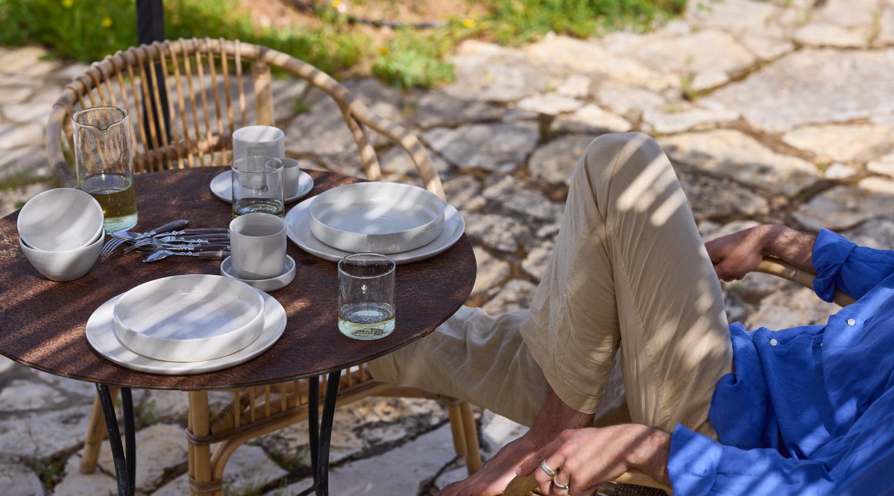 Ein runder Tisch im Freien ist gedeckt mit weißem Geschirr und gefüllten Gläsern. Daneben sitzt ein Mensch, zurückgelehnt in einem Stuhl.
