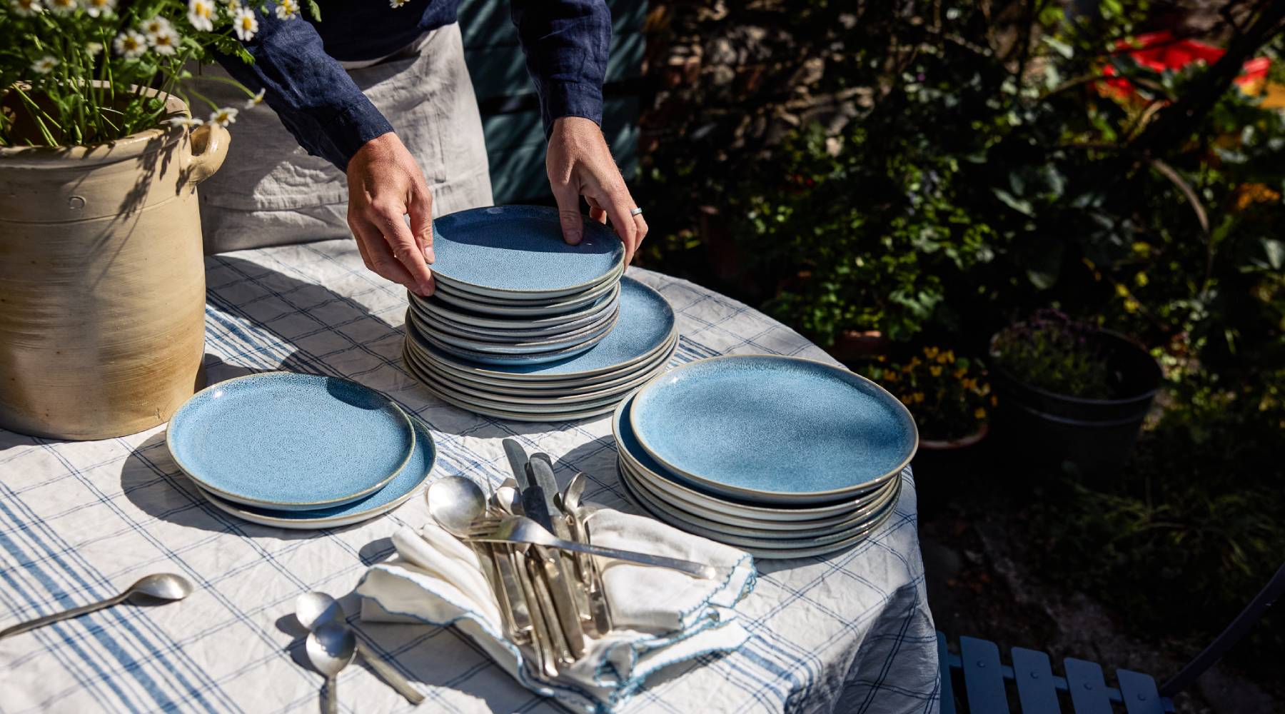 Ein runder Tisch im freien mit karierter Tischdecke. Darauf steht eine große Vase mit Blumen und blaue Teller sind gestapelt. Im Vordergrund liegt Besteck auf Stoff Servietten. Zwei Hände heben Teller von einem der Stapel an.