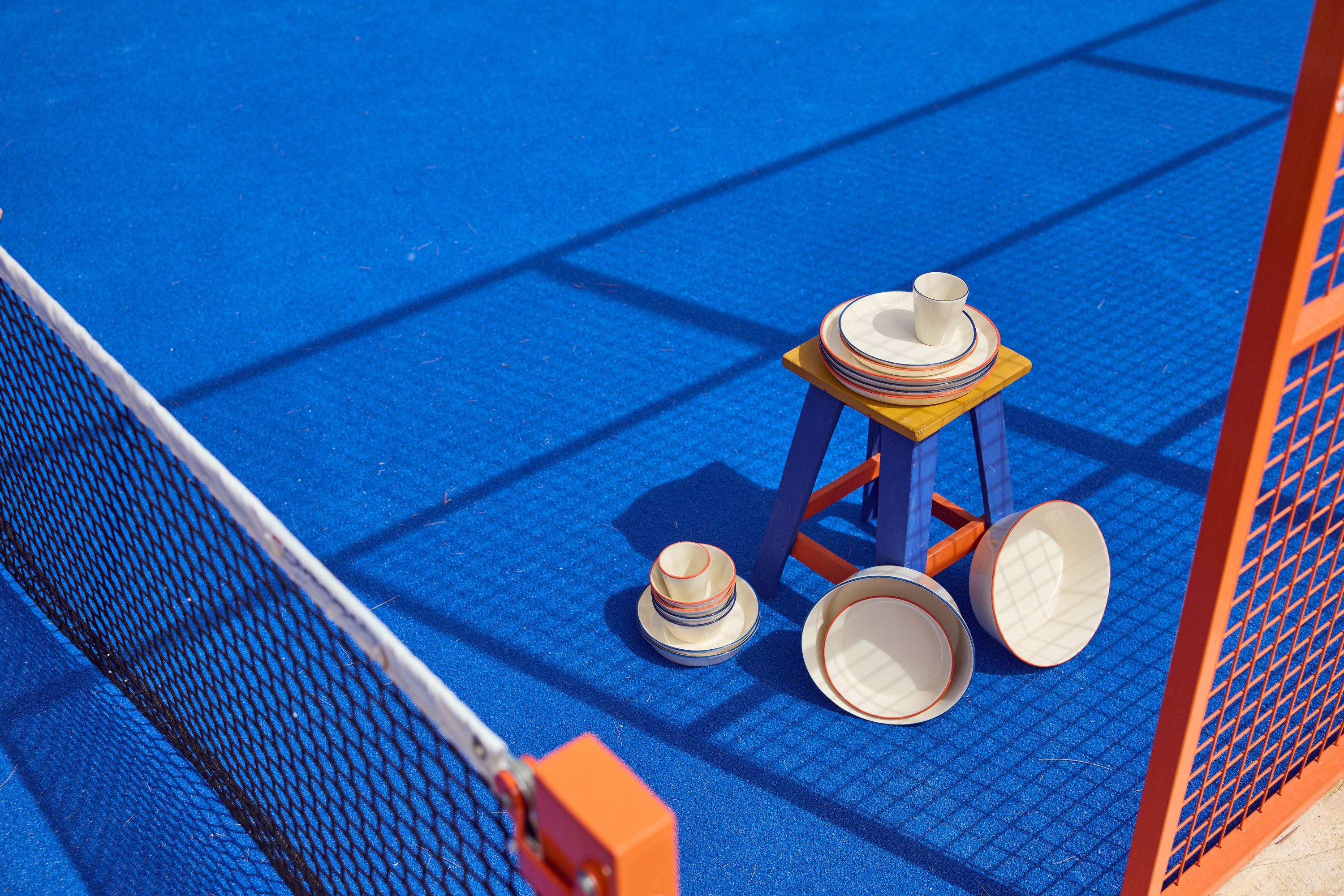 Keramik-Geschirrset der Kollektion Aberta auf einem blauen Padel-Court: Teller, Schalen und ein Becher mit orangefarbenen und blauen Randstreifen, arrangiert auf einem kleinen gelb-blauen Holzhocker, daneben weitere gestapelte Schalen auf dem Court-Boden. Im Vordergrund das Netz und ein orangefarbener Pfosten.