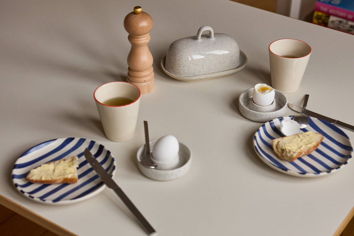 Gedeckter Frühstückstisch mit gesprenkelter Keramik-Butterdose, zwei Eierbechern mit Eiern, cremefarbenen Bechern mit rotem Rand und blau-weiß gestreiften Tellern.