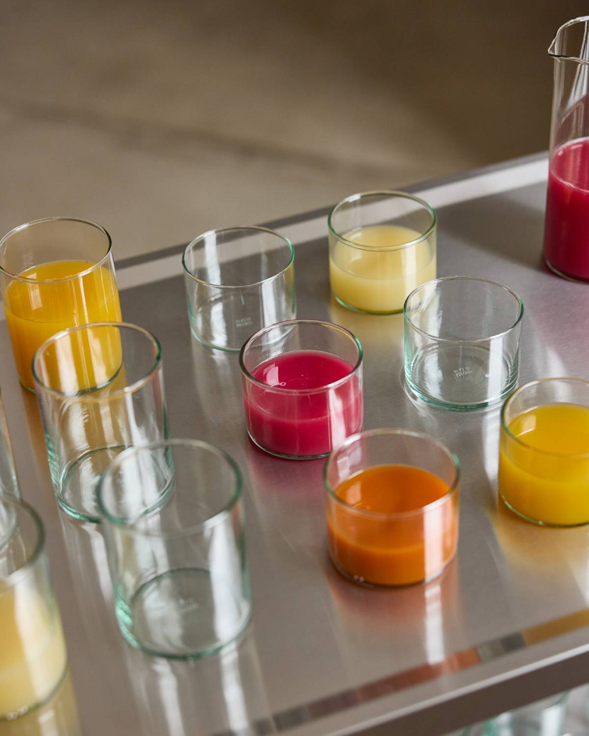 Gläser aus grünlichem Glas auf einem Edelstahlregal, teils leer, teils gefüllt mit Säften in Orange, Gelb und Pink.