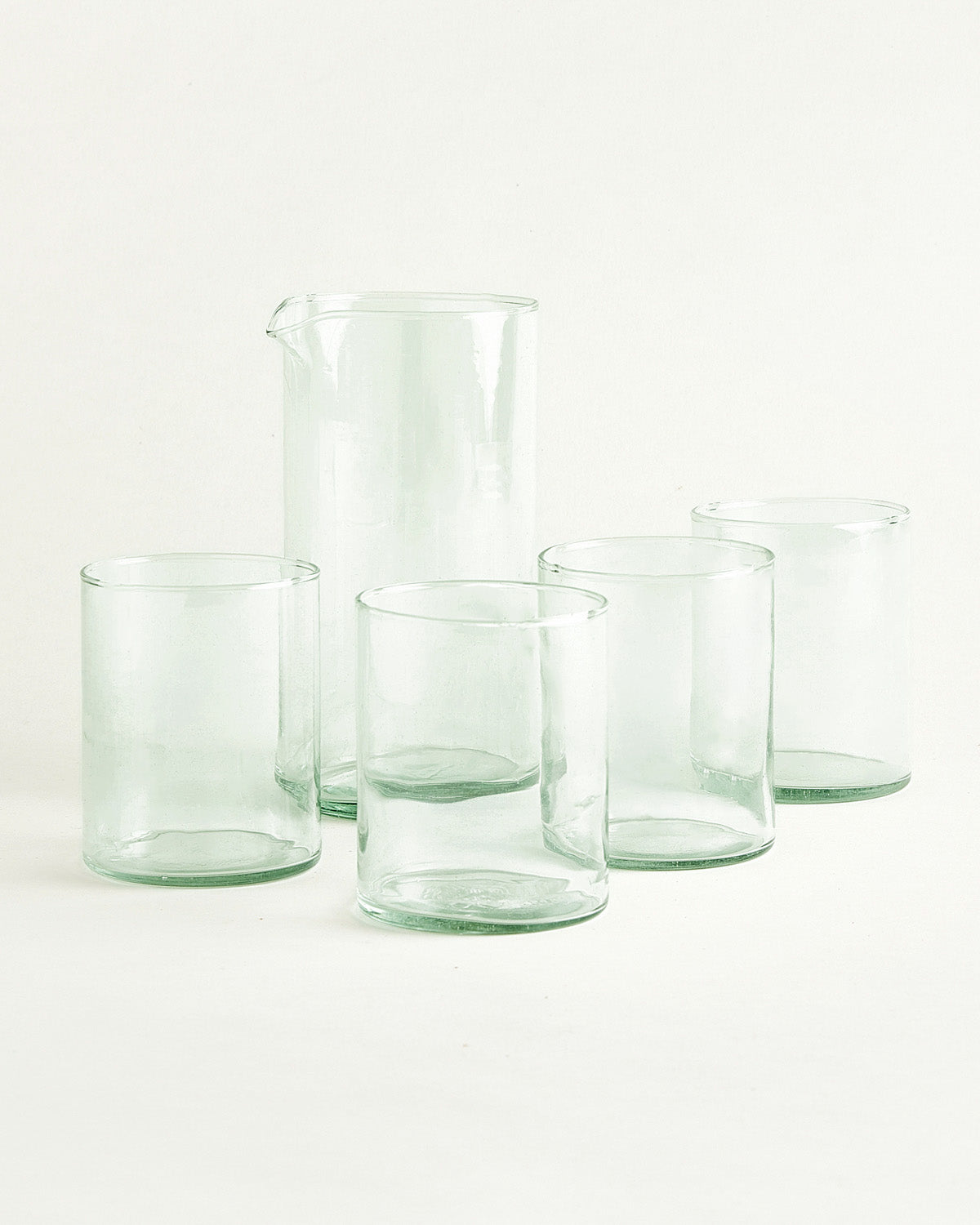 Vier hohe Trinkgläser und eine Karaffe aus grünlichem Glas vor hellem Hintergrund.