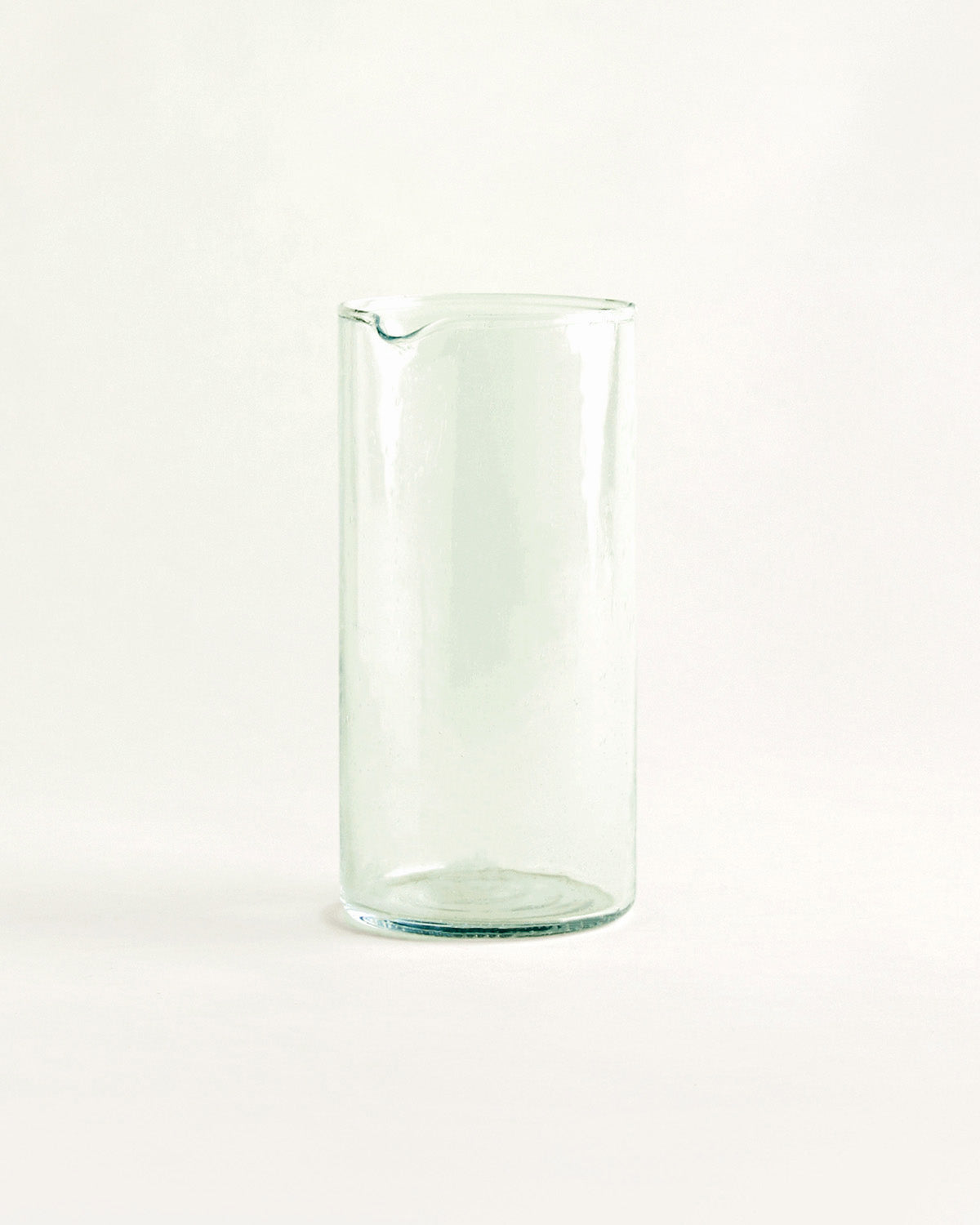 Schlanke, zylindrische Karaffe aus grünlichem Glas mit Ausguss, vor hellem Hintergrund.