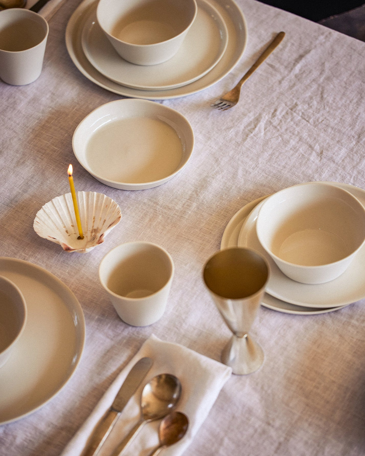 Gedeckter Tisch mit heller Leinentischdecke, cremefarbenem Keramikgeschirr, Bechern und Schalen, einer Muschel mit brennender Kerze, einem silbernen Kelch und Besteck auf einer Stoffserviette