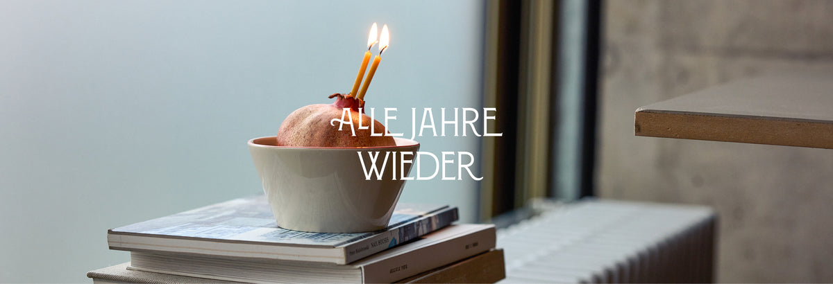 Alle Jahre wieder – ein Granatapfel mit zwei brennenden Kerzen in einer hellen Keramikschale auf einem Bücherstapel am Fenster