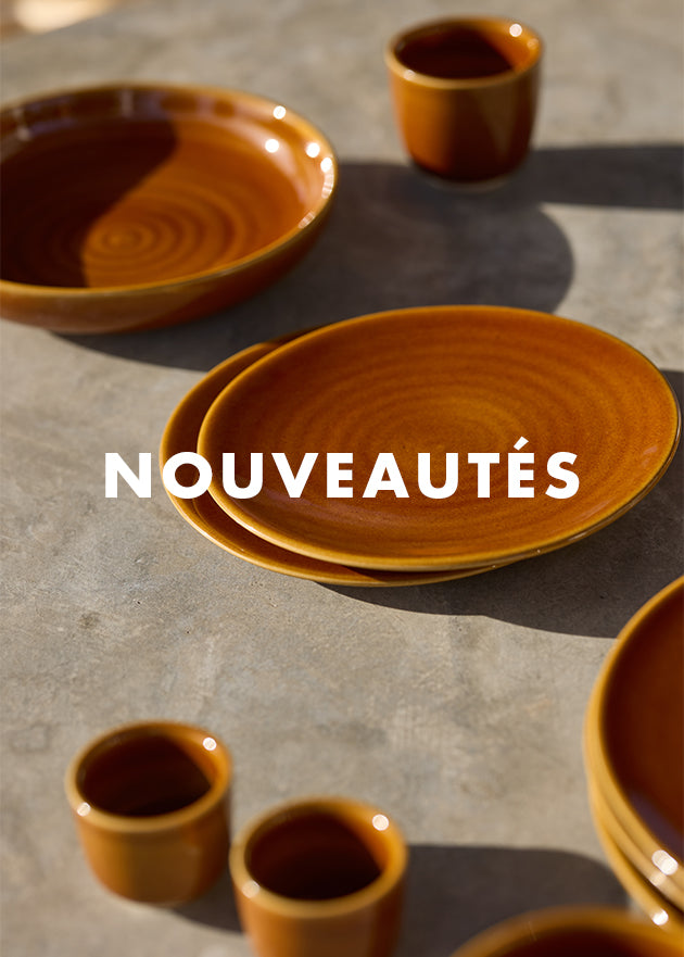 Close-up of onomao’s new amber-toned collection: flat plates, bowls, and small cups on a grey concrete surface. Centered text reads ‘NOUVEAUTÉS’ (New in the assortment).