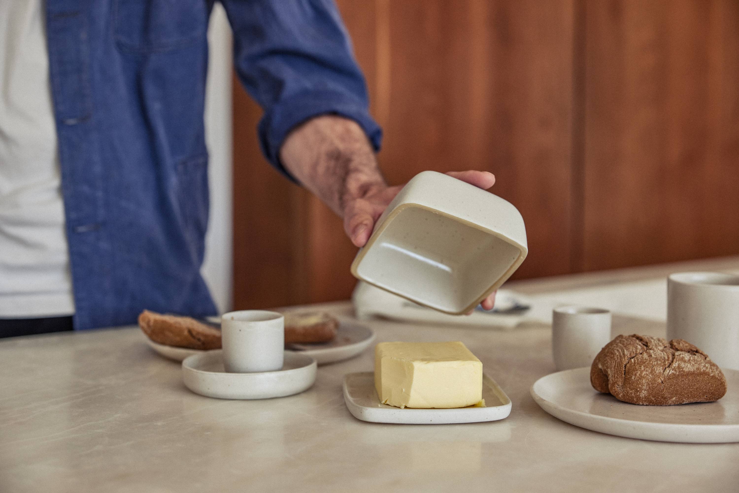Eine Person in blauer Jeansjacke hebt den Deckel einer cremefarbenen Keramik-Butterdose an. Auf einem kleinen Keramikteller darunter liegt ein Block gelber Butter. Im Hintergrund sind weitere Keramikbecher und ein Teller mit einem Stück Roggenbrot auf einer hellen Steinoberfläche zu sehen.