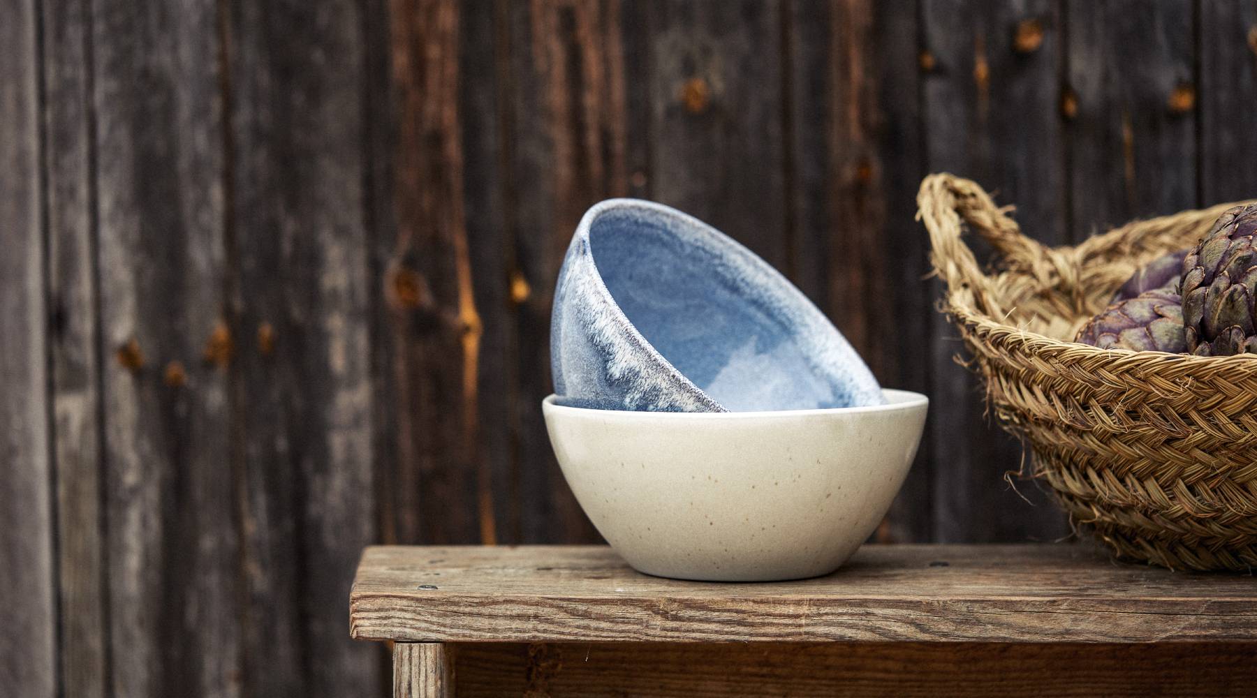 Zwei Keramikschalen stehen auf einem rustikalen Holztisch vor einer dunklen Holzfassade. Die untere Schale ist cremefarben, die obere zeigt innen eine blaue, verlaufende Glasur. Daneben steht ein geflochtener Korb mit violetten Artischocken.