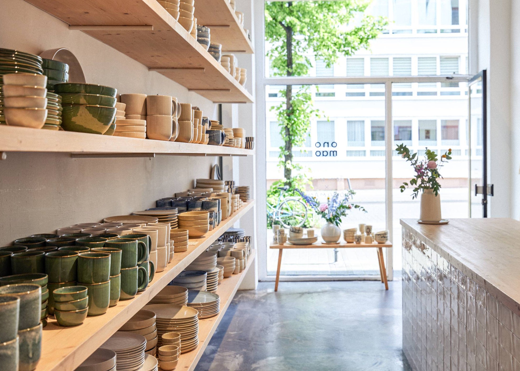 Helles Keramikgeschäft mit Holzregalen voller Tassen, Schüsseln und Teller in Grün- und Beigetönen, mit Blick auf eine sonnige Straße durch eine geöffnete Glastür.