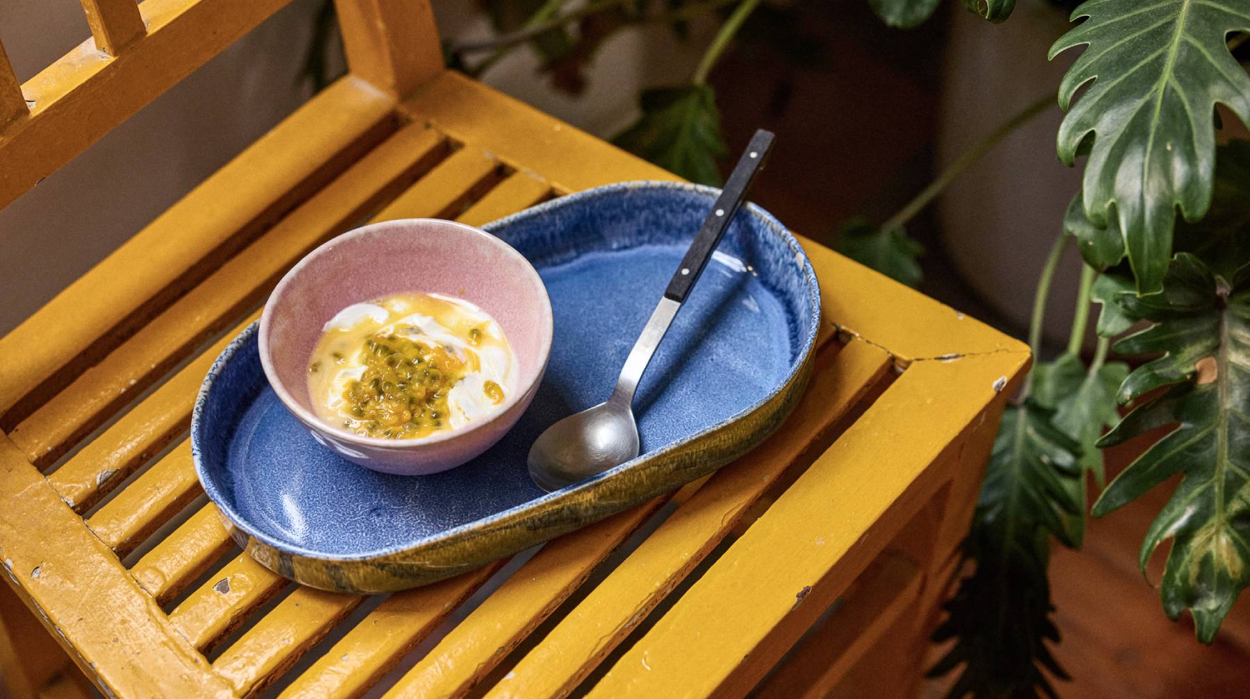 Roséfarbene Keramikschale mit Joghurt und Passionsfruchtpüree auf einem größeren blauen Keramikteller, der auf einer gelben Holzbank steht; im Hintergrund rechts eine grüne Zimmerpflanze. Weißer Schriftzug: ‘MÜSLISCHALEN’.