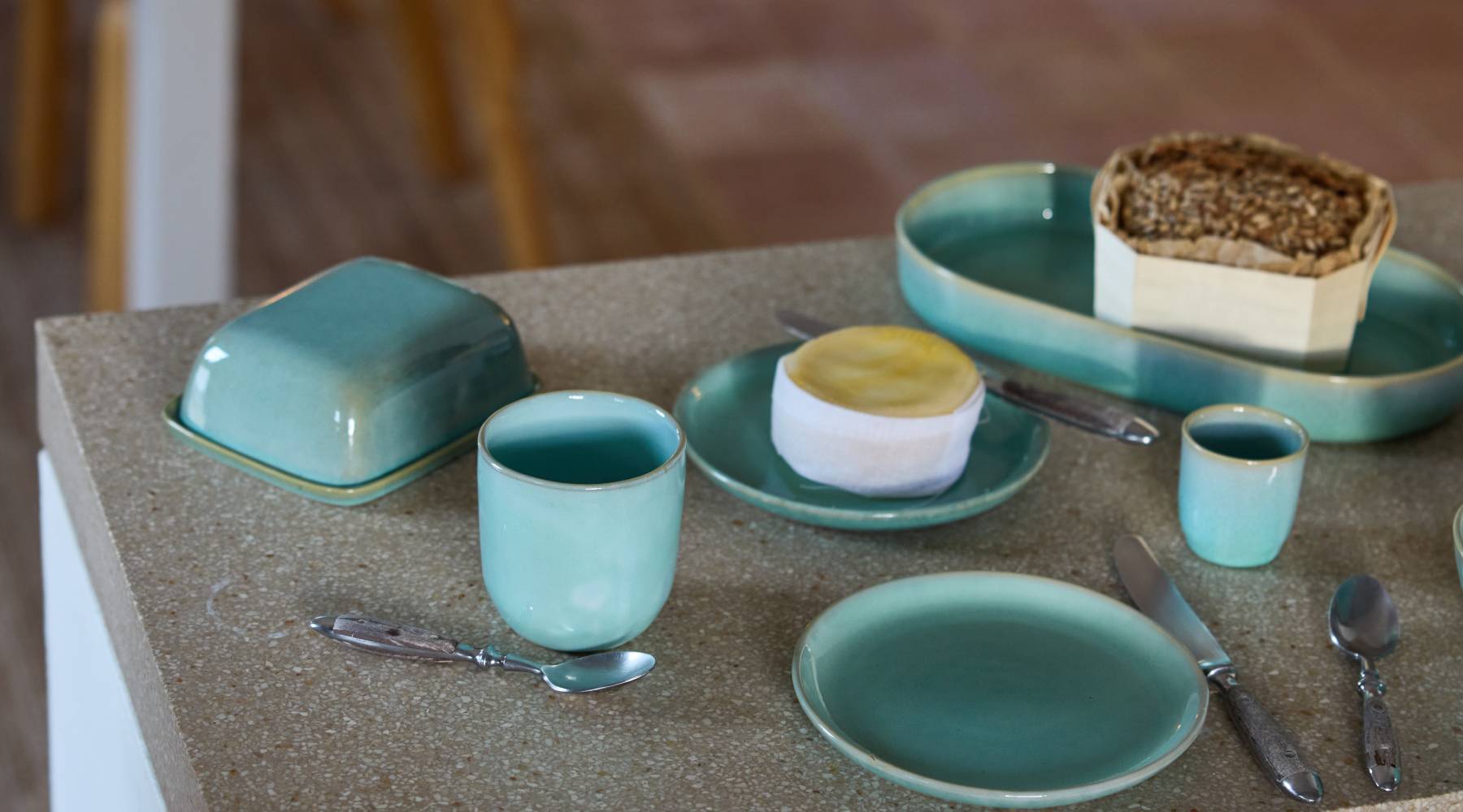 Auf einem hellen Terrazzo-Tisch arrangiertes Frühstücksgeschirr in Türkistönen: links eine geschlossene Butterglocke, davor eine Tasse mit kleinem Löffel, rechts ein kleiner Teller mit rundem Weichkäse, daneben ein Messer und ein weiterer Löffel. Im Hintergrund ein längliches Schälchen mit einem Brotlaib und ein winziger Espresso-Becher.