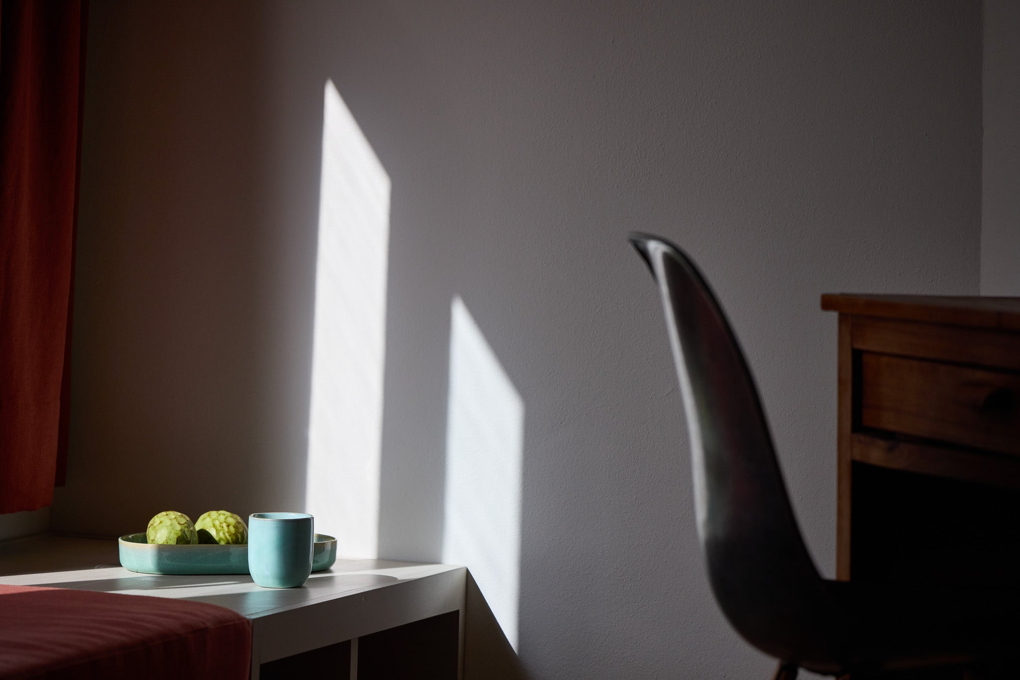 Helles Morgenlicht wirft zwei rechteckige Schatten an eine weiße Wand. Im Vordergrund steht ein weißer Tisch mit einer türkisfarbenen Keramikschale gefüllt mit grünen Früchten und einem passenden Keramikbecher. Im Hintergrund sind ein roter Vorhang und ein Holzschrank zu sehen.