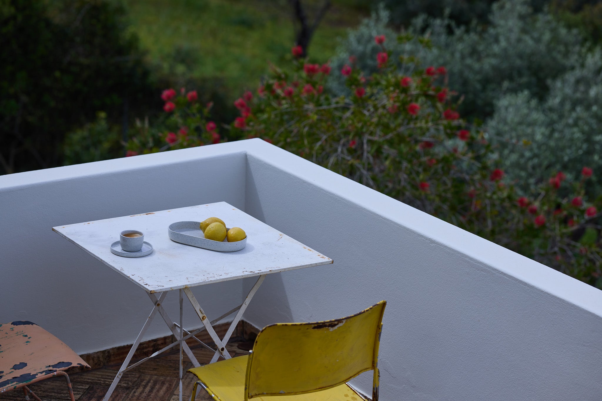 Weißer Vintage-Klapptisch auf einer mediterranen Terrasse mit einem hellgrauen Keramikbecher und einem grauen Tablett mit gelben Zitronen. Daneben ein gelber Metallstuhl, im Hintergrund rote Blüten und grünes Gebüsch.