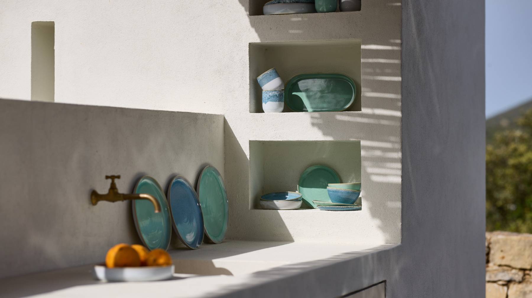 Eine Weiße Küchenzeile im Freien mit Wasserhahn. Im Vordergrund steht ein Teller mit Orangen. Dahinter sind drei Teller an die Wand gelehnt. Im Regal steht weiteres Türkis und Blau gefärbtes Geschirr. 