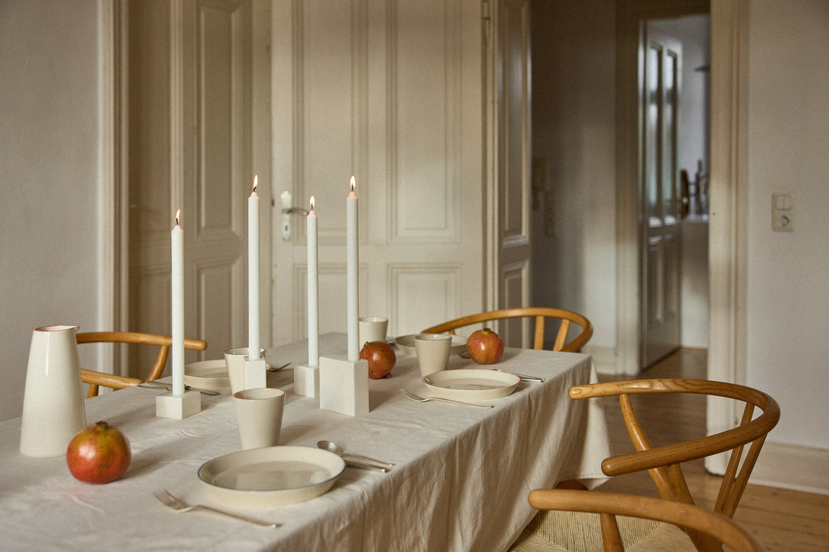 Festlich gedeckter Tisch mit heller Leinentischdecke, Keramiktellern und Bechern, weißen Kerzen in eckigen Kerzenhaltern und Granatäpfeln in einem hellen Altbauzimmer
