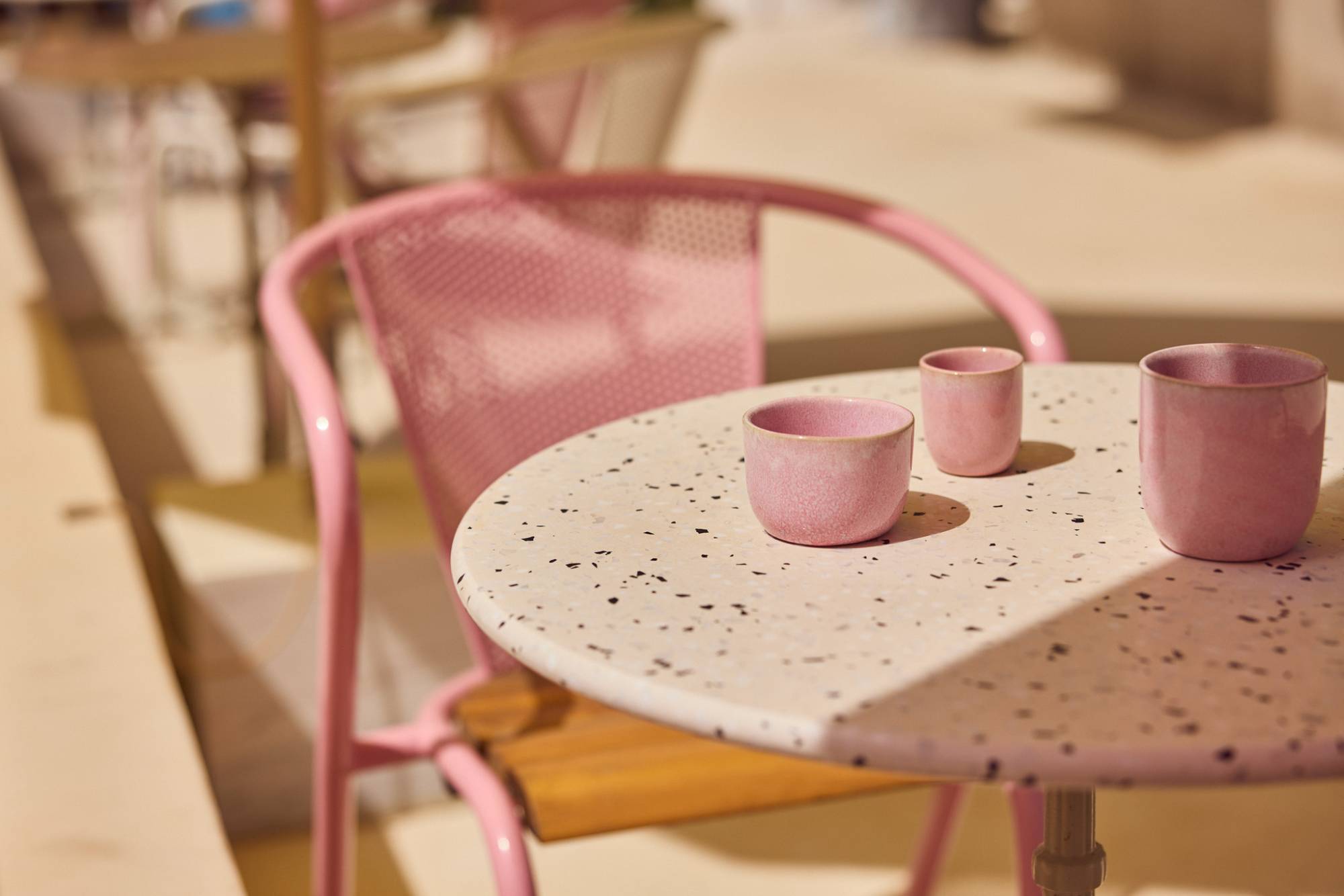 Zwei rosafarbene Keramiktassen auf einem weißen Terrazzo-Tisch mit schwarzen Sprenkeln, daneben ein rosafarbener Metallstuhl im französischen Bistro-Stil. Im Hintergrund weitere Tische und Stühle, leicht unscharf.
