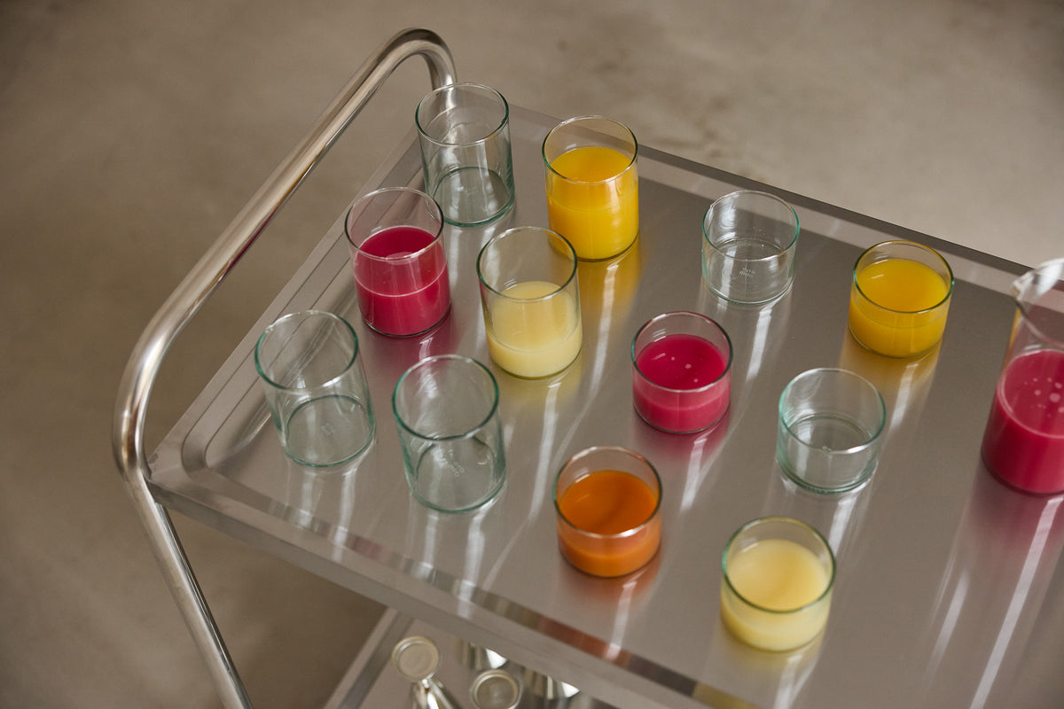 Mundgeblasene KLAR-Gläser auf einem verchromten Servierwagen, teils leer mit grünlichem Schimmer, teils gefüllt mit Orangensaft, Bananensaft und rotem Fruchtsaft.