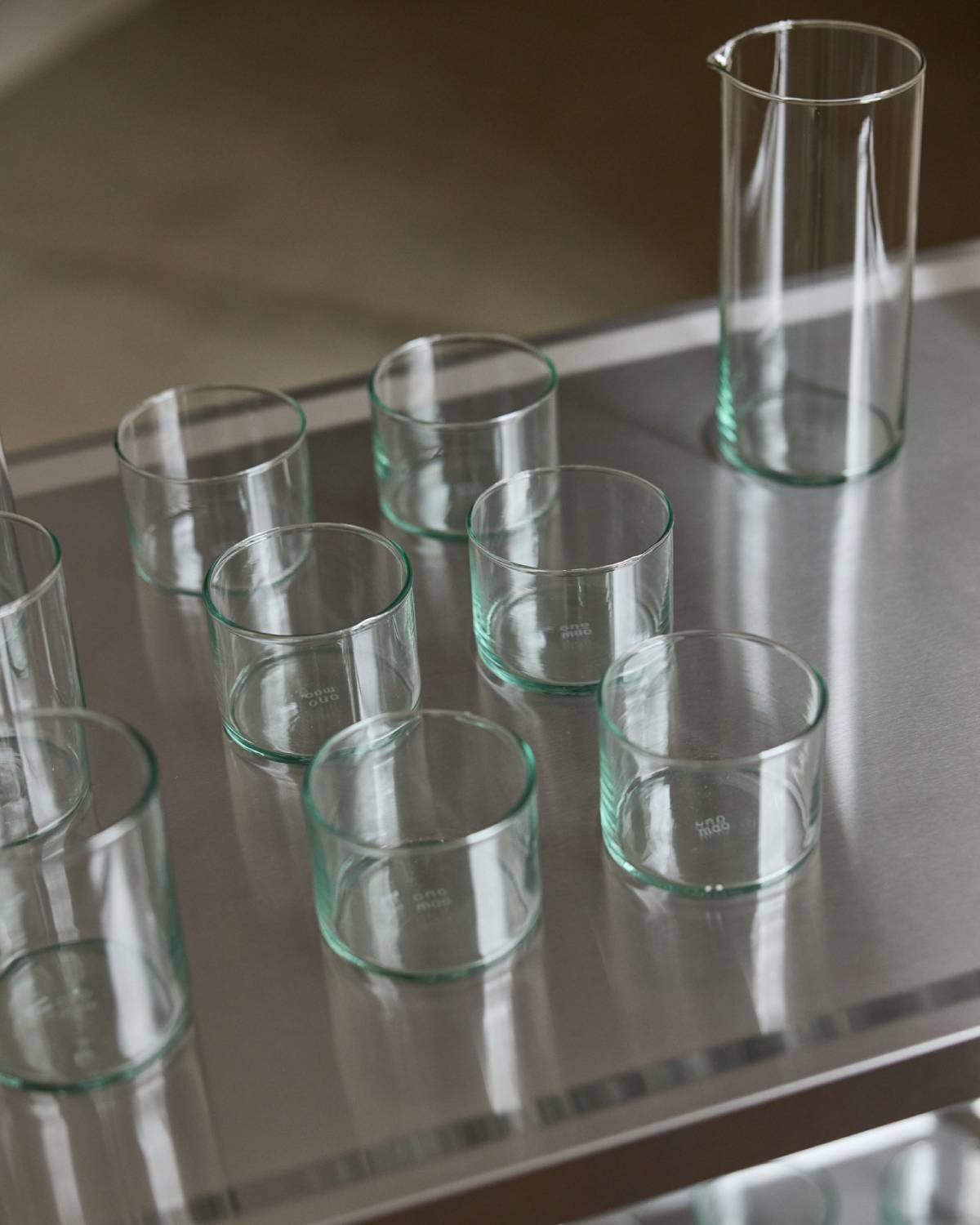 Sieben Trinkgläser und eine Karaffe aus leicht grünlichem Glas mit onomao-Prägung, aufgereiht auf einem Edelstahlregal.