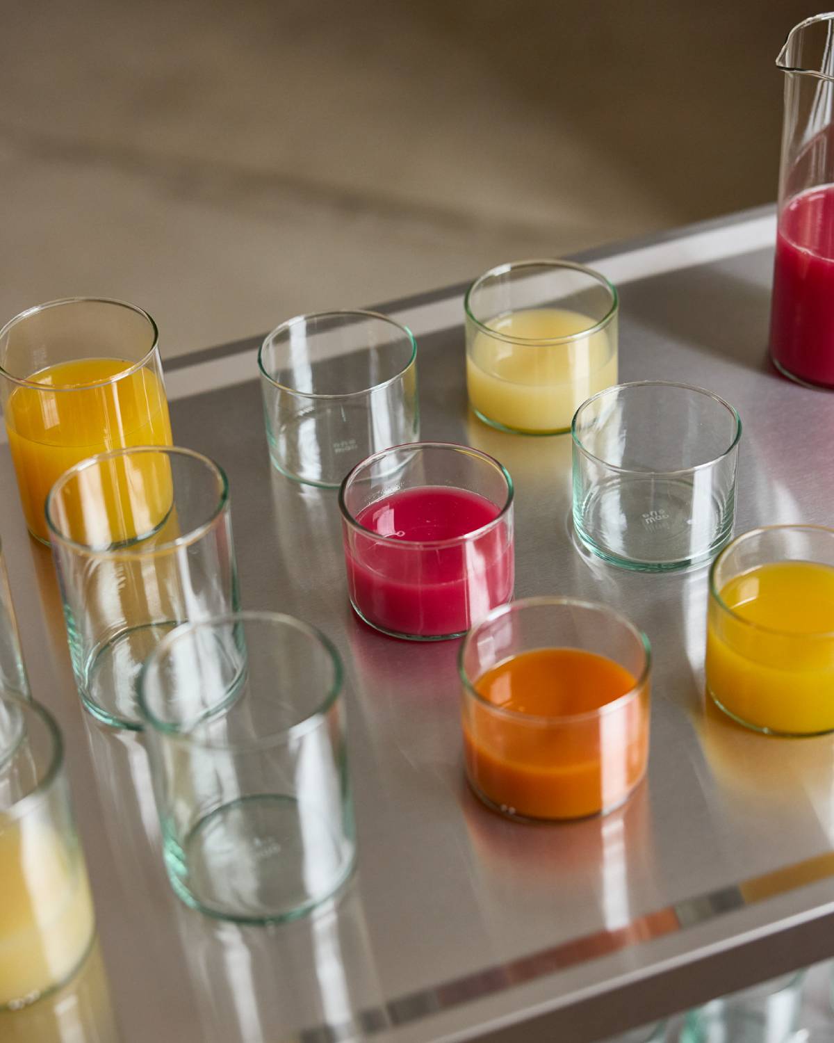 Hohe und niedrige Gläser aus grünlichem Glas auf einem Edelstahlwagen, teils leer, teils gefüllt mit Säften in Gelb, Orange und Pink.