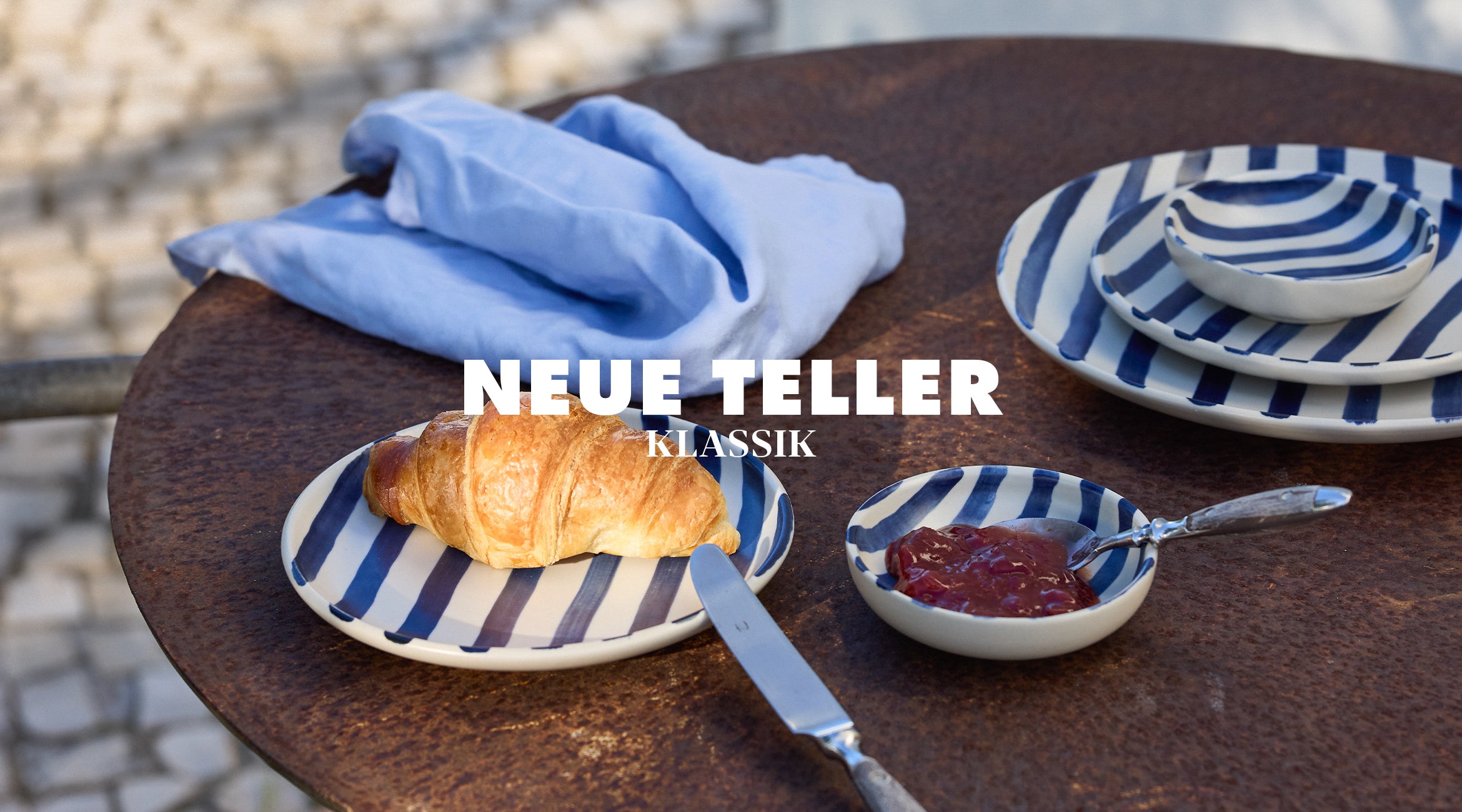 Neue Teller Klassik von onomao: Frühstücksszene auf einem runden Bistrotisch mit blau-weiß gestreiften Steingut-Tellern, einem Croissant, einer Schale mit Marmelade und einer hellblauen Leinenserviette im Hintergrund.