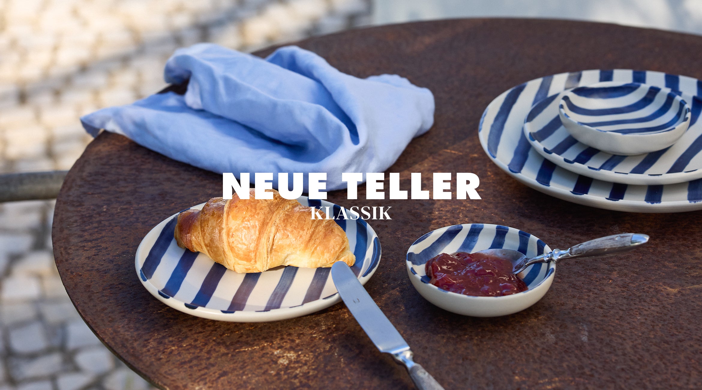 Neue Teller Klassik von onomao: Frühstücksszene auf einem runden Bistrotisch mit blau-weiß gestreiften Steingut-Tellern, einem Croissant, einer Schale mit Marmelade und einer hellblauen Leinenserviette im Hintergrund.