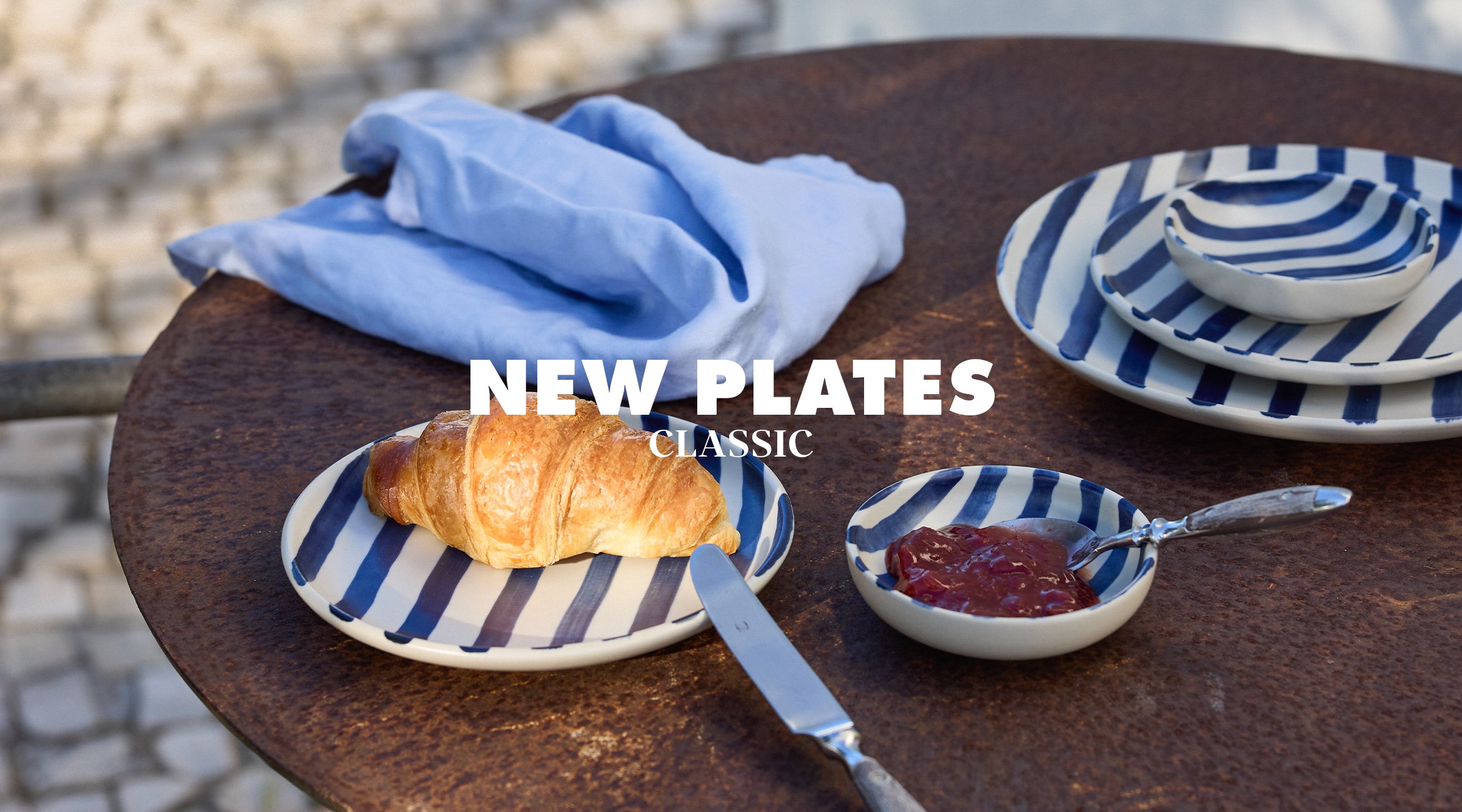 New Plates Klassik by onomao: Breakfast scene on a round bistro table with blue and white striped stoneware plates, a croissant, a small bowl of jam and a light blue linen napkin in the background.